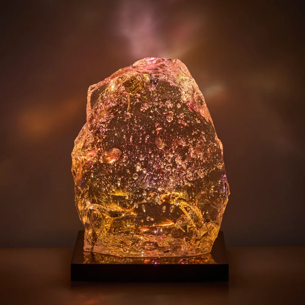 Glowing glass sculpture CF5, No Right Angle series, amber tones with dense sparkling inclusions and a soft pink glow near the tip, raw natural form on a hand-crafted base containing LED lighting